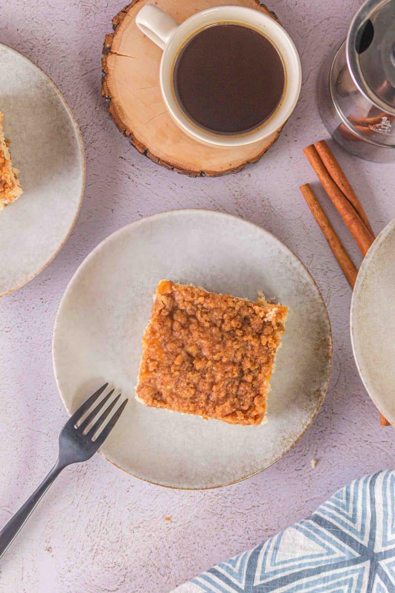 Top down more distant image of a piece of coffee cake on a saucer with a cup of coffee nearby and cinnamon sticks for ambience.