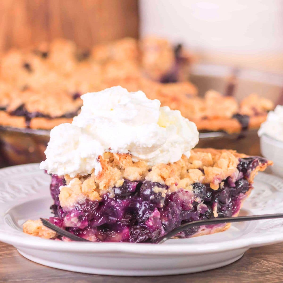 A tableview of a slice of blueberry pie crumble with the first bite sitting on a fork - Hostess At Heart