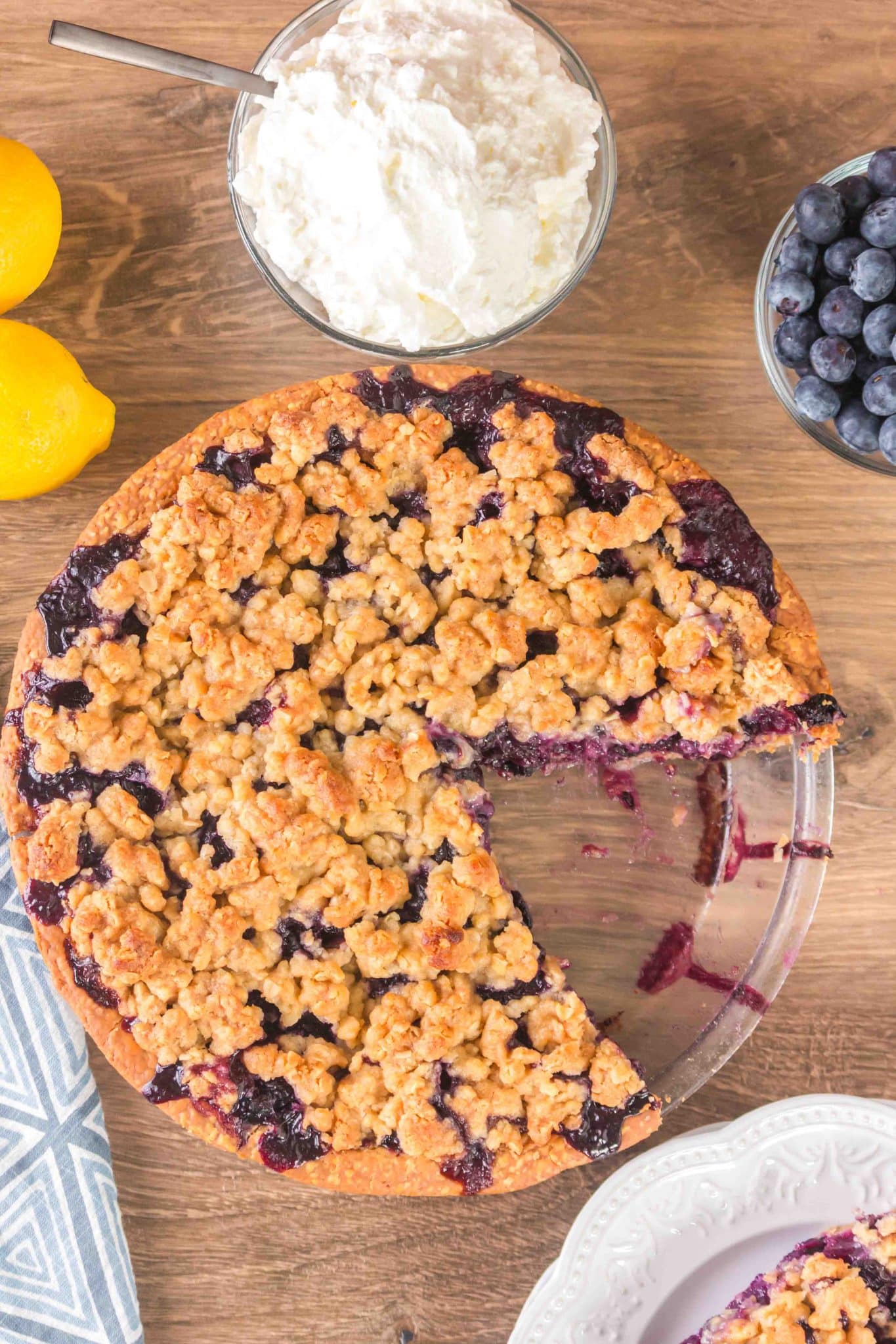 Top down view of a blueberry pie with crumble topping with the first slice removed to a serving plate - Hostess At Heart