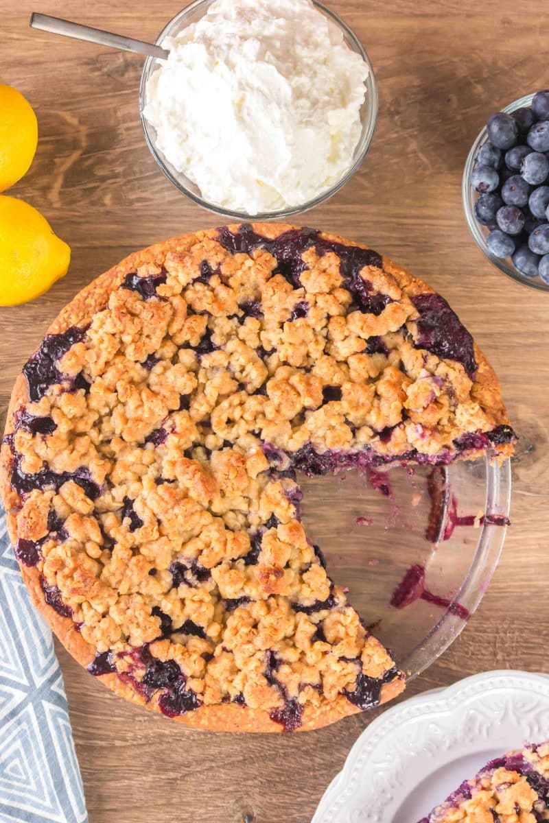 Top down view of a blueberry pie with crumble topping with the first slice removed to a serving plate - Hostess At Heart