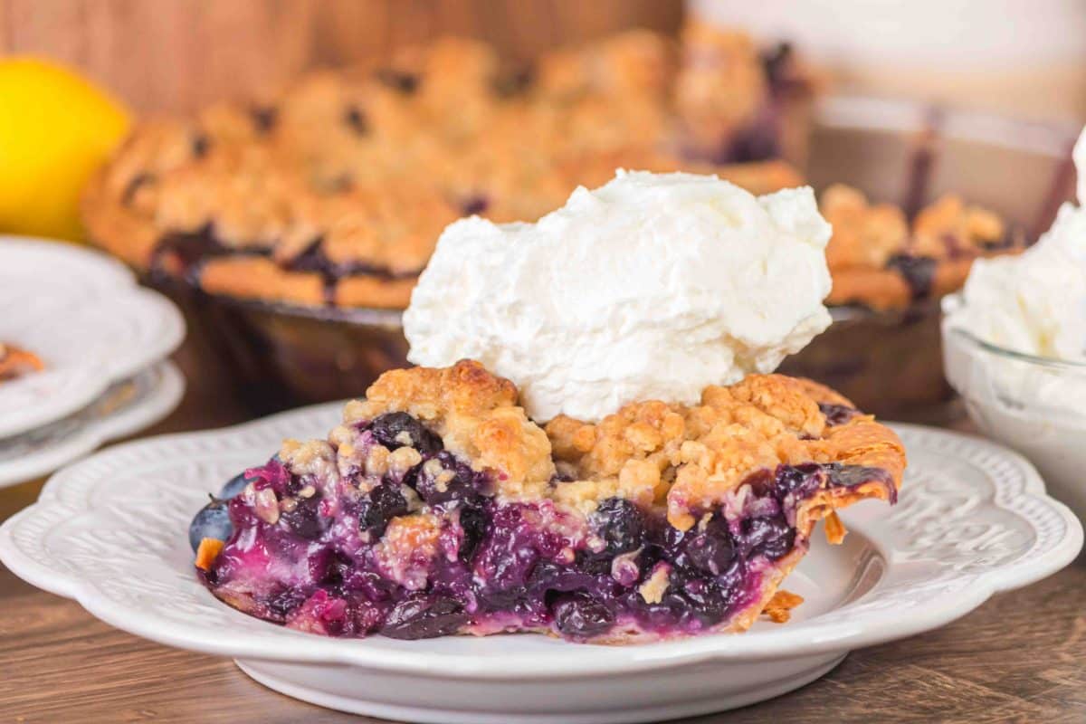 Sideview of a slice of blueberry crumble pie topped with whipped cream - Hostess At Heart