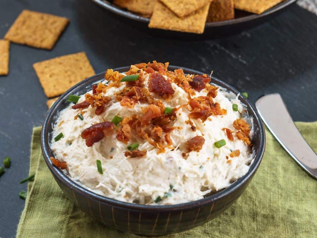 Angled view of a bowl of cold and creamy crab dip topped with crumbled bacon and freshly chopped chives - Hostess At Heart