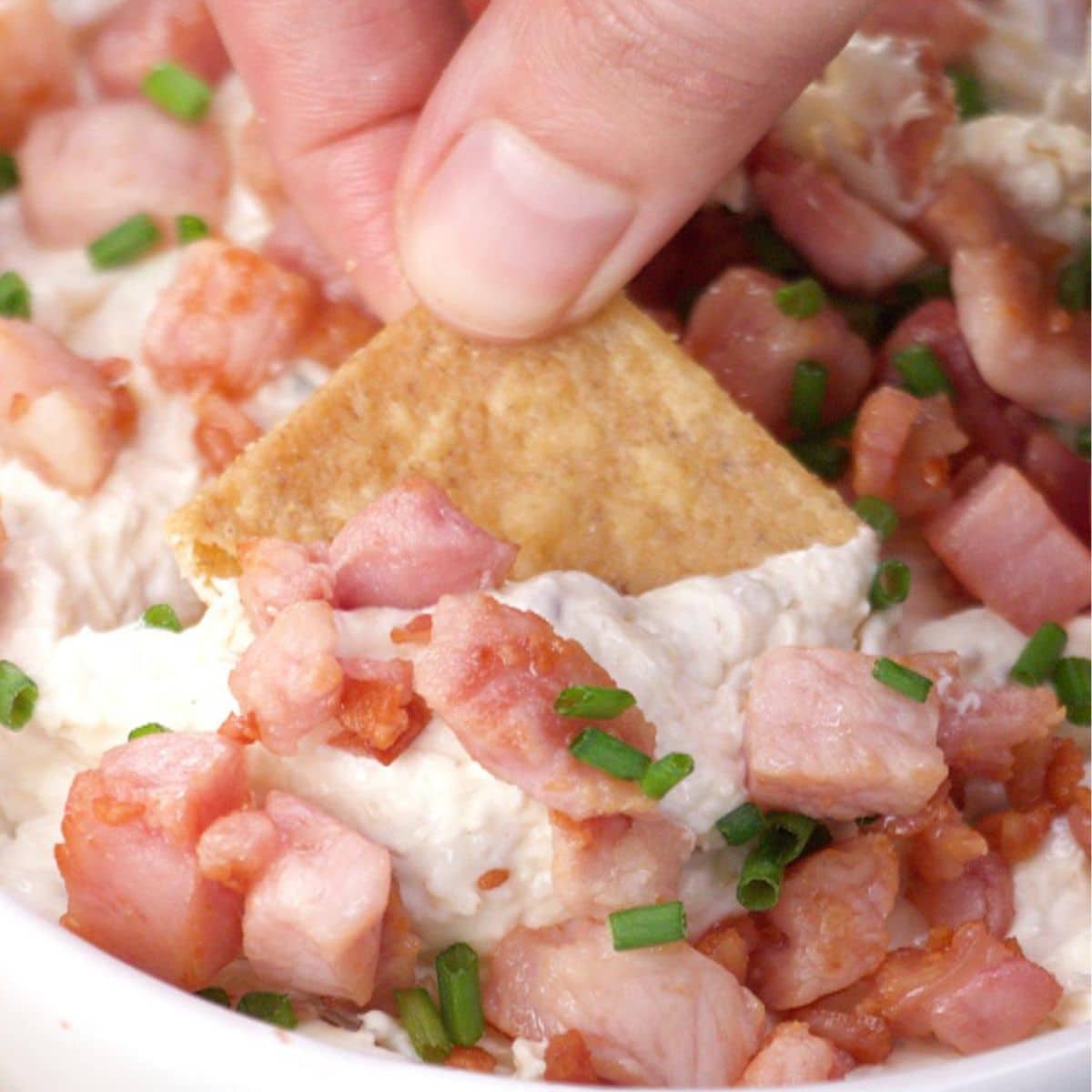 Fingers holding a wheat cracker filled with cold creamy crab dip topped with thick cooked bacon pieces - Hostess At Heart