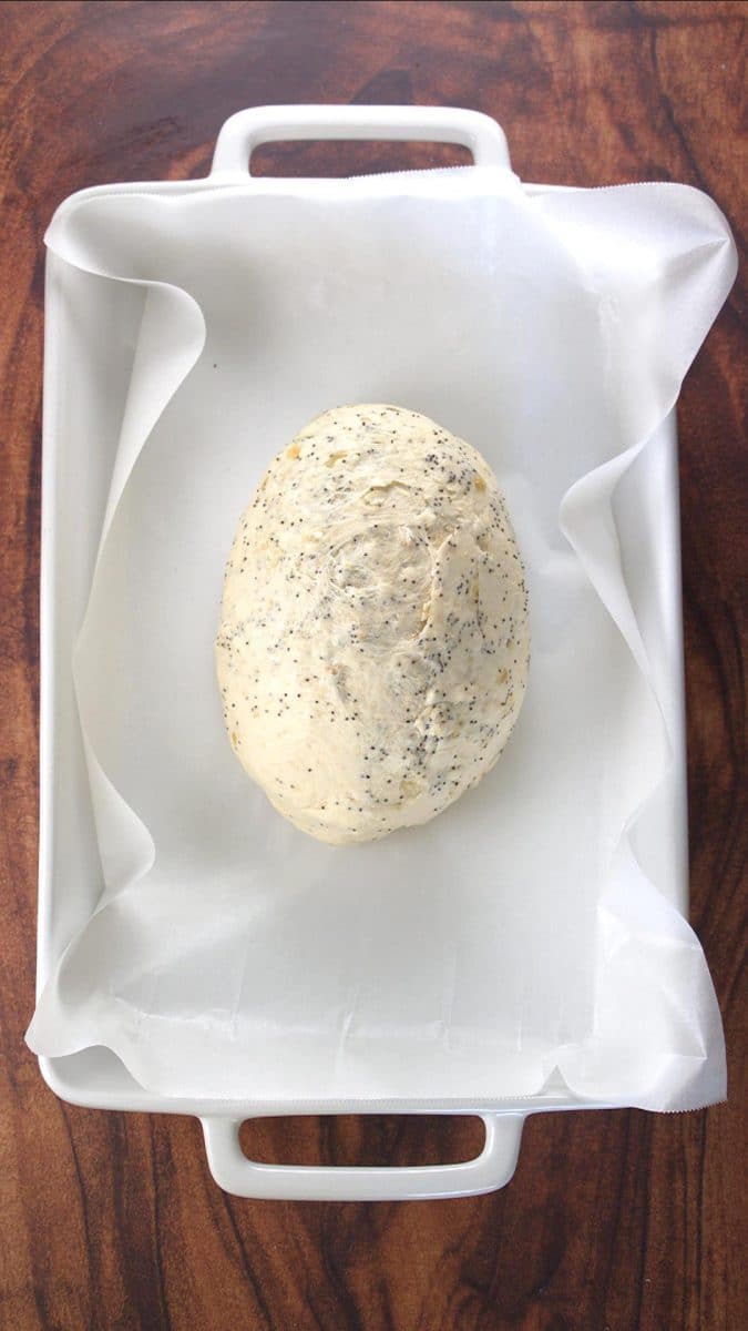 A shaped loaf of seeded bread dough sitting in a parchment paper-lined baking dish - Hostess At Heart