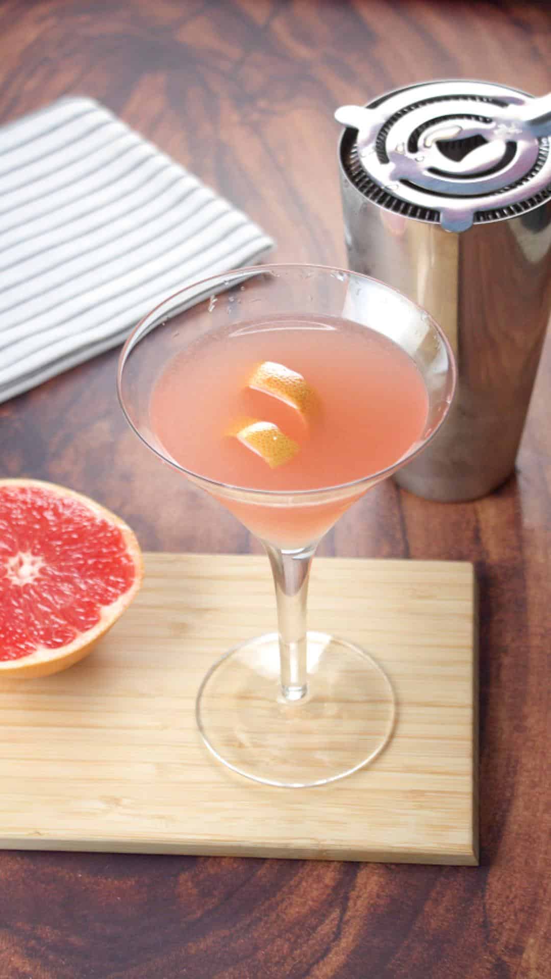 Sideview of a fresh red grapefruit martini setting next to a shaker and a cut pink grapefruit - Hostess At Heart