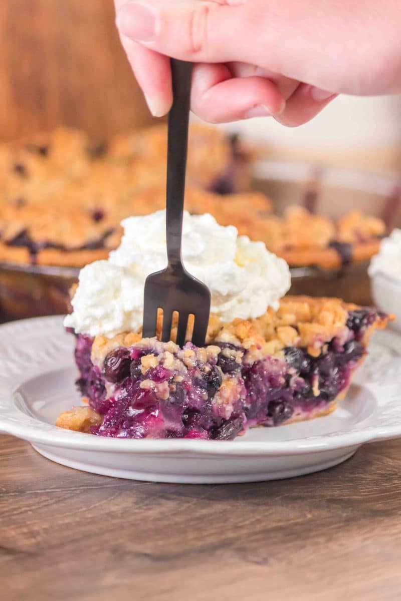 A fork removing the tip of a slice of blueberry pie with crumble topping and topped with whipped topping - Hostess At Heart
