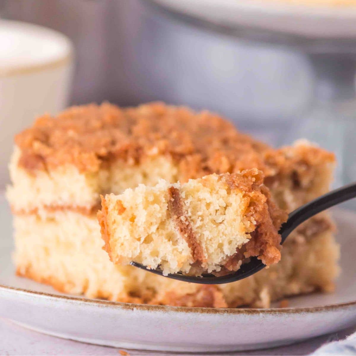 A fork holding a bite of sourdough discard coffee cake showing a cinnamon ribbon running through it. A slice of the sourdough cake sits behind it on a plate - Hostess At Heart