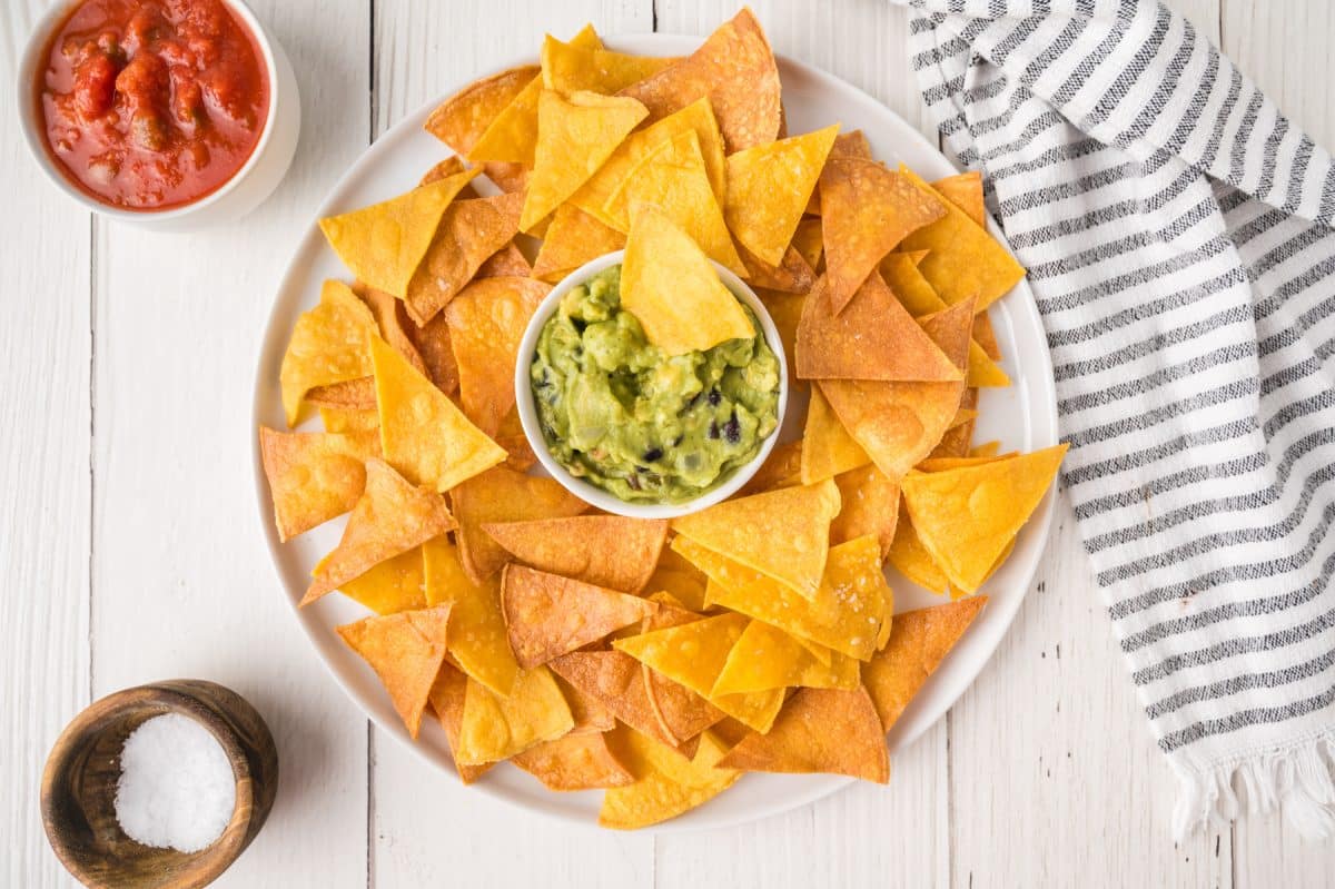 Top down view of a white platter containing a bowl of guacamole surrounded by homemade baked corn tortilla chips - Hostess At Heart