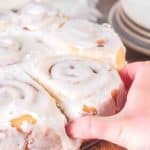 A hand that's reaching into some baked and frosted sourdough cinnamon rolls and pulling one out - Hostess At Heart