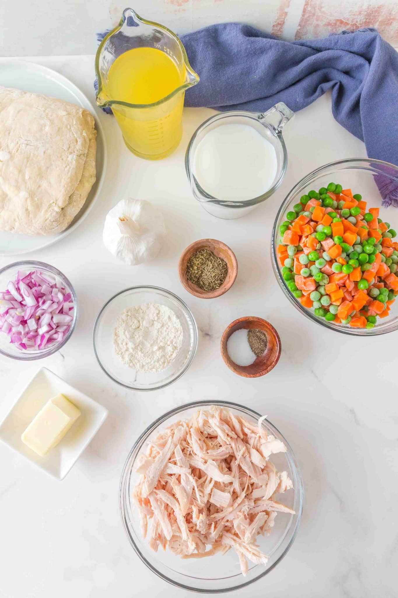 Ingredients used to make chicken biscuit casserole including shredded chicken, frozen peas and carrots, broth, seasonings, milk, onion, butter, flour, garlic, and biscuit dough - Hostess At Heart