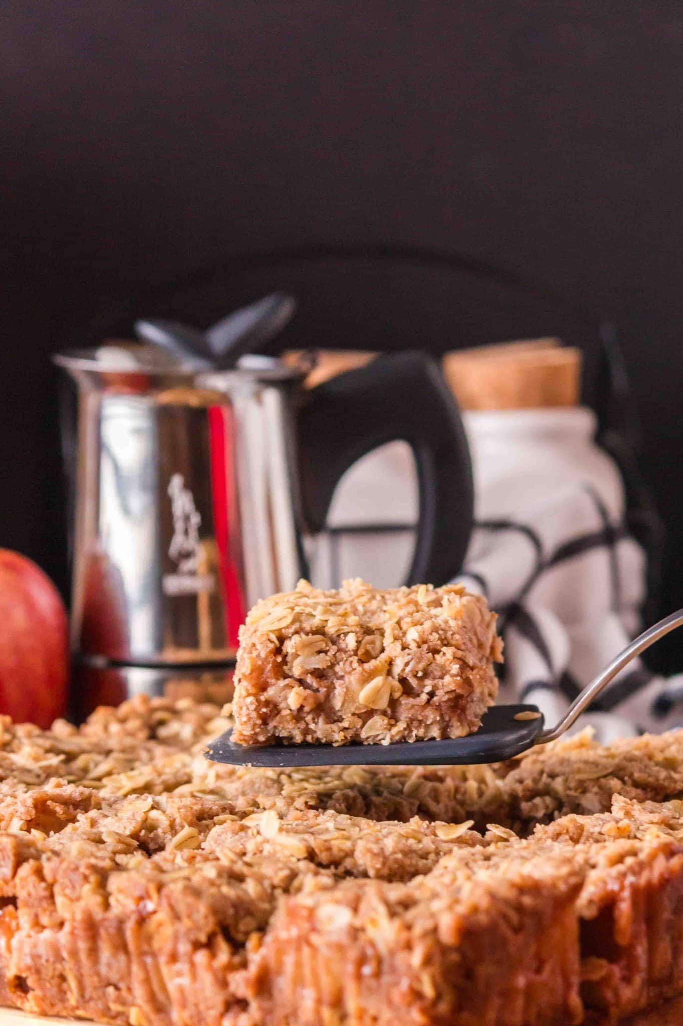 Sideview of a apple cinnamon crumble bar sitting on a spatula hovering over the rest of the bars - Hostess At Heart