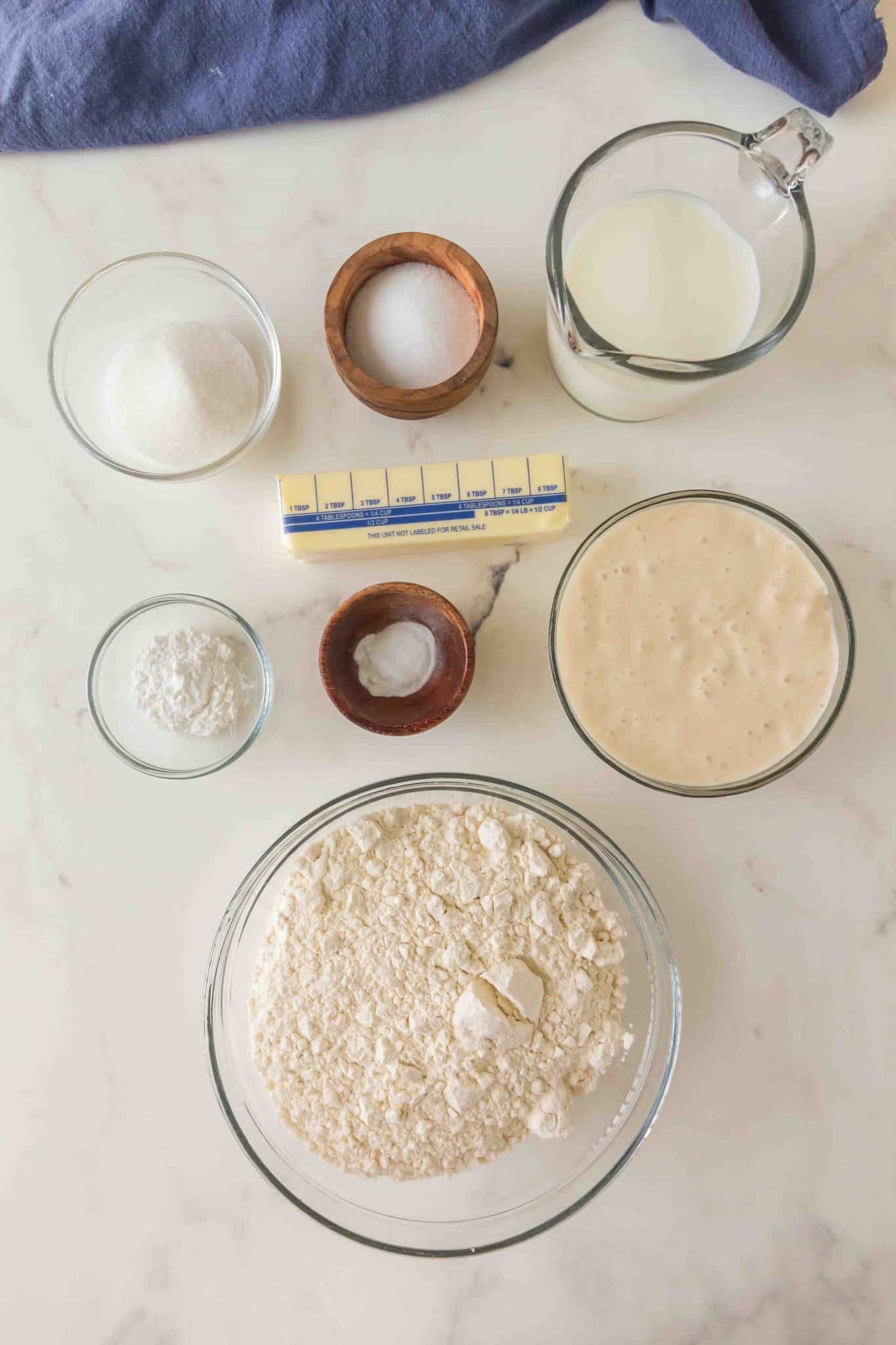 Top down view of the ingredients necessary to make sourdough starter biscuits including flour, sugar, butter, baking powder, baking soda, salt, milk, and sourdough starter discard. Hostess At Heart