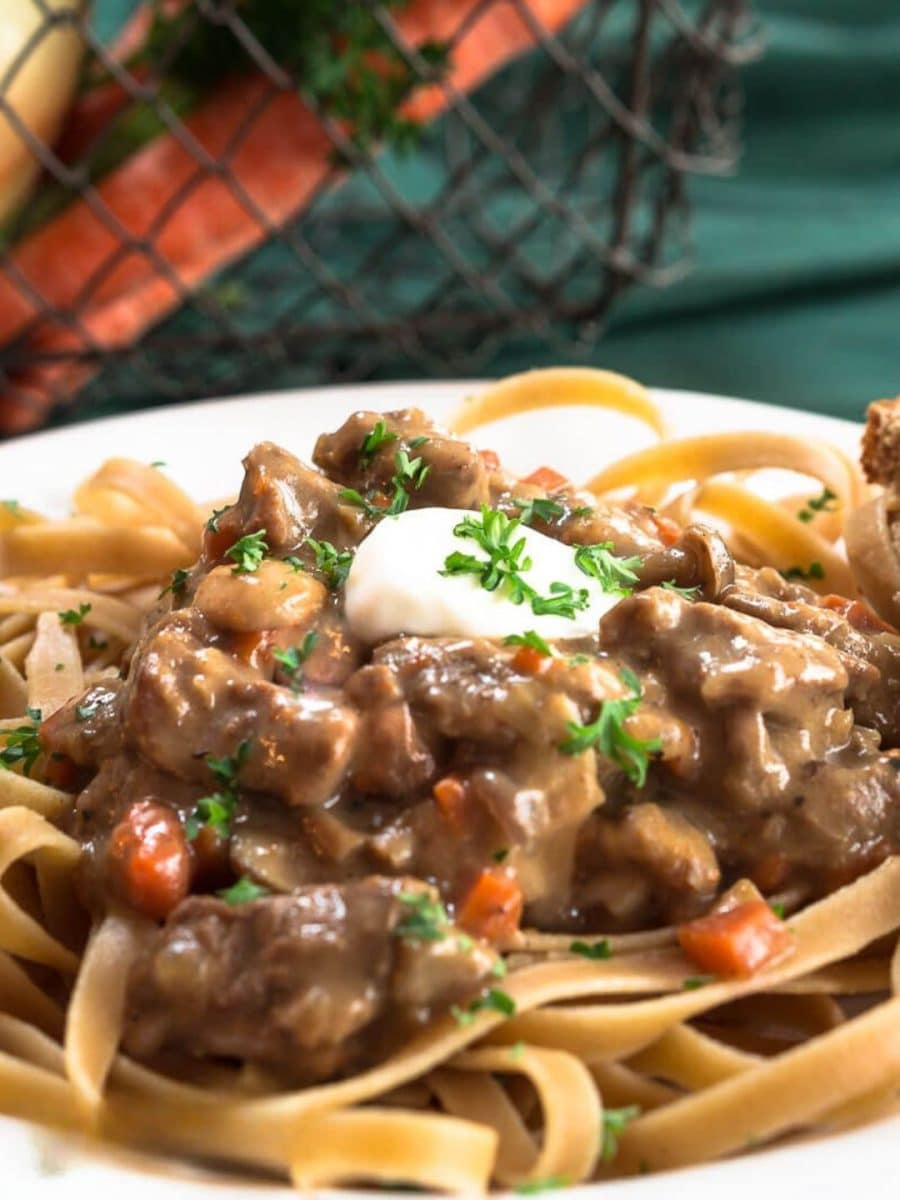 Close up view of tender chunks of beef in a stroganoff sauce served over whole grain pasta noodles and garnished with sour cream and chopped parsley - Hostess At Heart