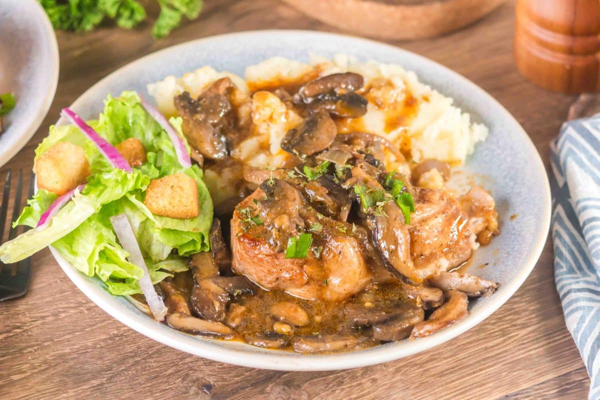 A dinner plate filled with a pork chop and mashed potatoes covered with a onion gravy. A side salad sits to the side - Hostess At Heart