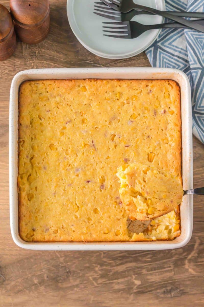 A baking dish filled with a holiday corn casserole baked golden brown. A serving spoon has scooped out a serving from the corner. Hostess At Heart