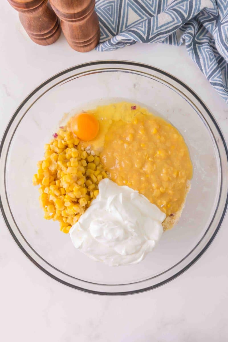 A mixing bowl filled with sour cream, corn, corn muffin mix, chopped onion, and an egg. Hostess At Heart