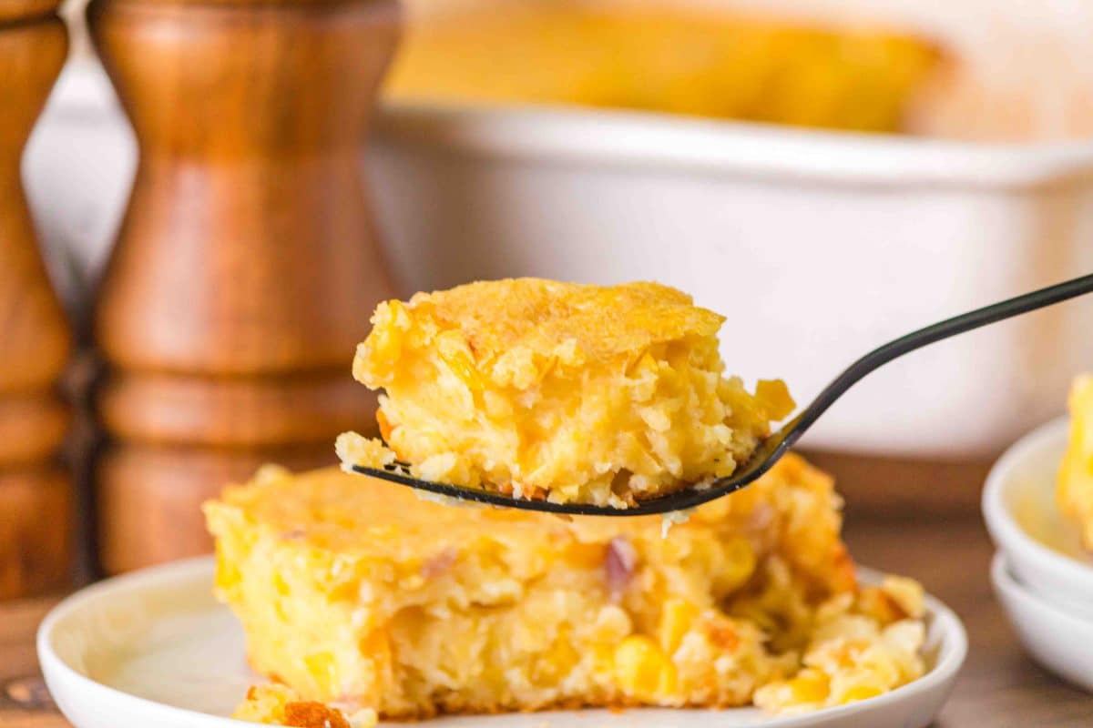 Sideview of a plate holding a serving of scalloped corn casserole. A fork hovers over the serving holding a thick moist bite. Hostess At Heart