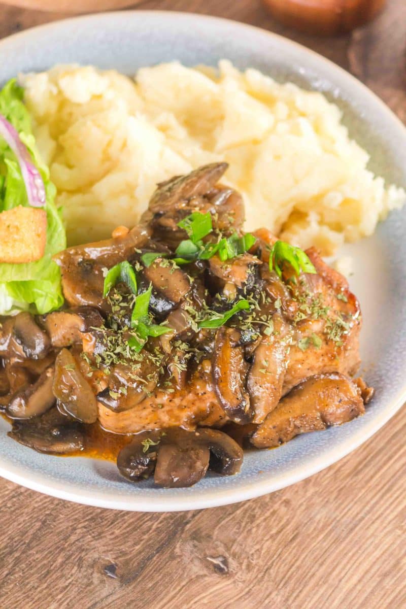 A sideview of a plate filled with a loaded pork loin chop in mushroom gravy. Hostess At Heart