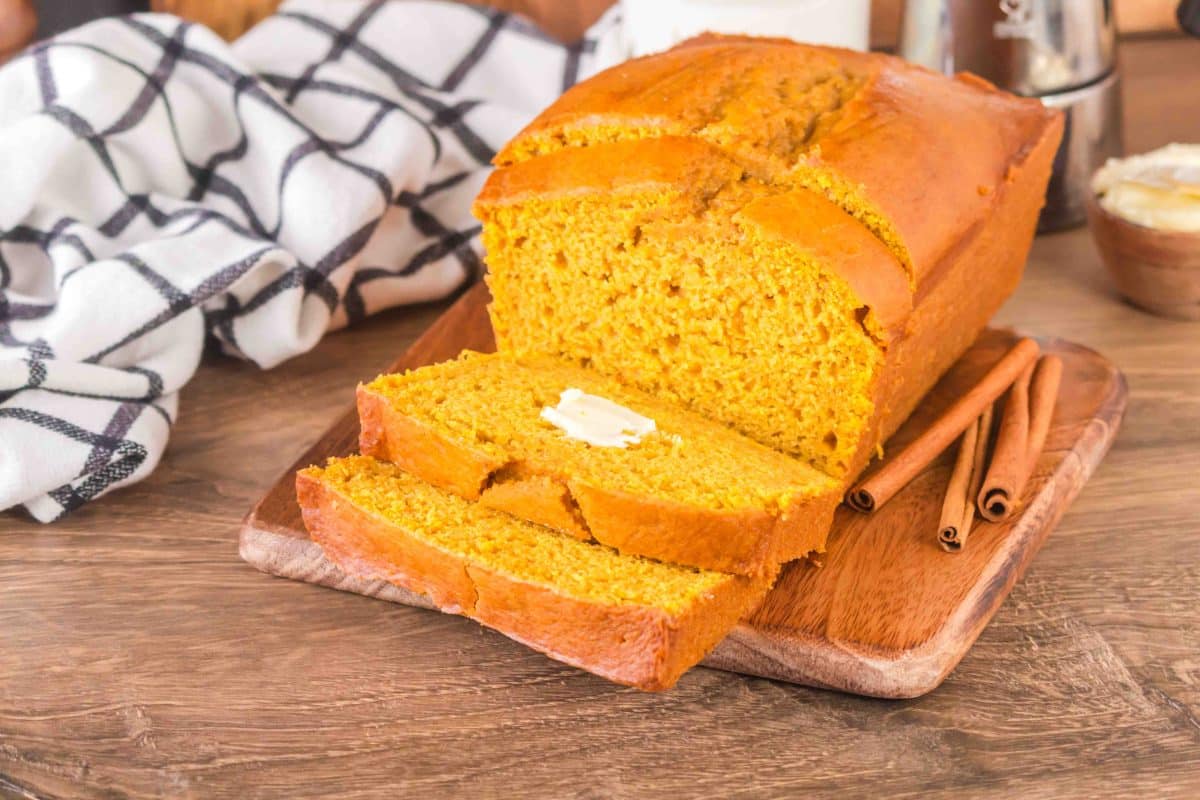 A loaf of sourdough pumpkin bread with the front slices fanned in front of the loaf. The top slice has a pad of butter on it. Hostess At Heart
