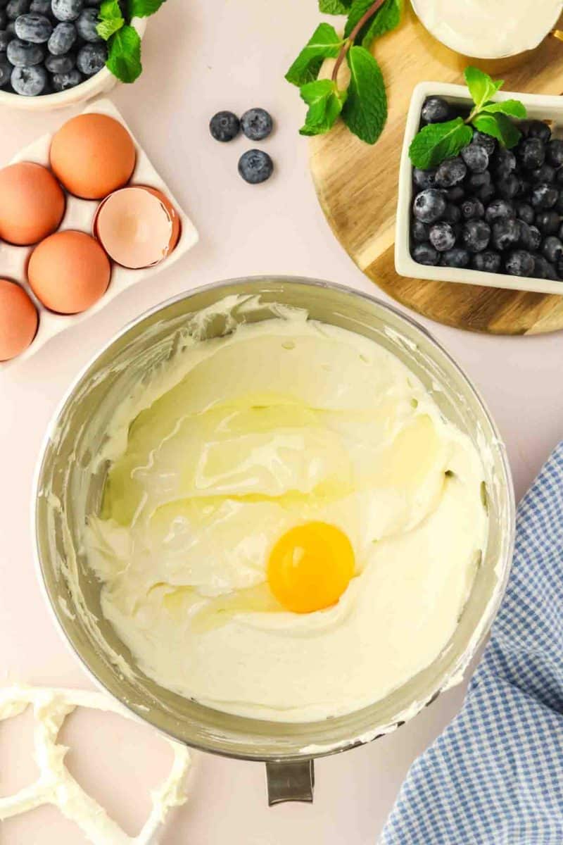 A whole egg added to a bowl of creamy cheesecake batter - Hostess At Heart