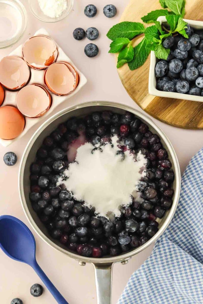Fresh blueberries cooking in saucepan with sugar and lemon juice for homemade blueberry cheesecake topping - Hostess At Heart