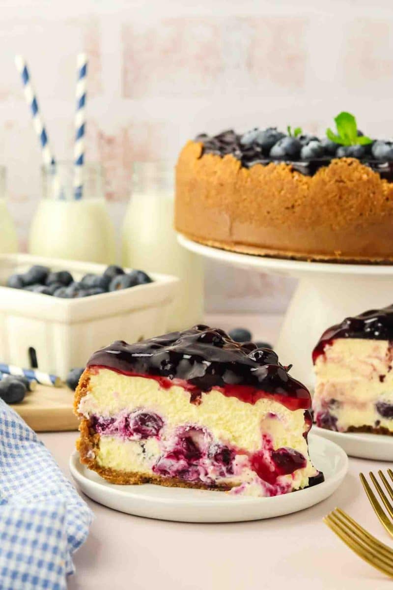 Side view of a creamy slice of blueberry cheesecake on a white plate showing graham cracker crust and blueberry-studded filling - Hostess At Heart