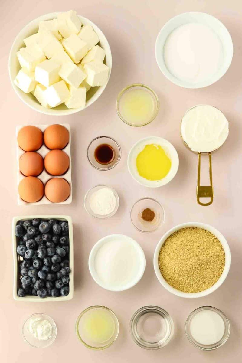Ingredients for blueberry cheesecake including cream cheese, eggs, fresh blueberries, graham cracker crumbs, cinnamon, corn starch, lemon juice, sugar, sour cream, vanilla extract, flour, water, and butter on pink background - Hostess At Heart