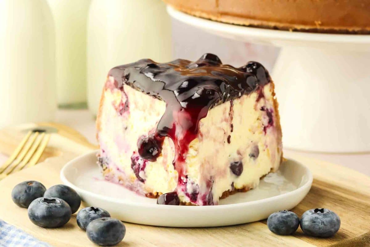 Front view of a slice of homemade blueberry cheesecake with fresh blueberries and glossy blueberry topping dripping down the side - Hostess At Heart