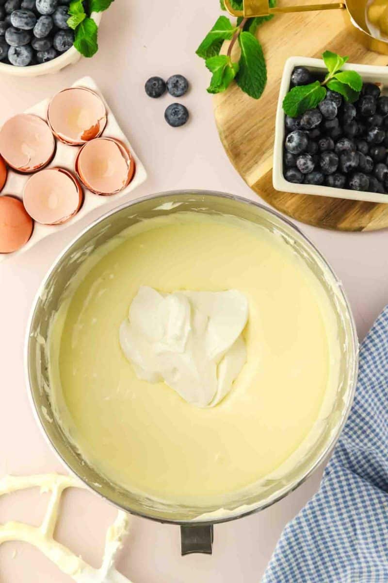 A dollop of sour cream added to the bowl of an electric mixer filled with creamy cheesecake filling - Hostess At Heart