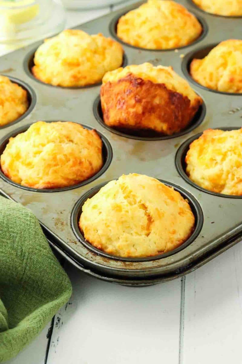 Golden brown cheddar cheese muffins in a muffin tin fresh from the oven. The Center one is tipped out showing the golden brown exterior - Hostess At Heart