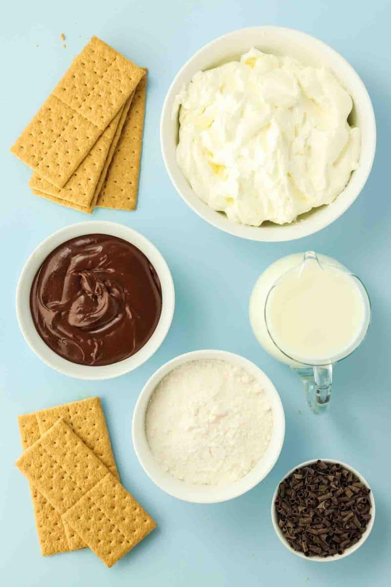 Ingredients for no-bake chocolate eclair cake including graham crackers, pudding mix, milk, whipped topping, and chocolate frosting, chocolate curls