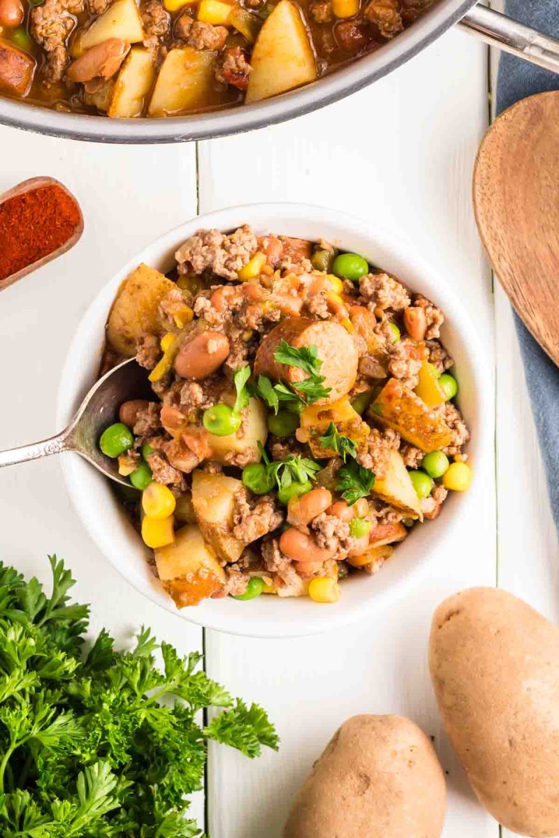Top down view of cowboy stew in a white bowl filled with ground beef, sausage, potatoes, corn, peas, and beans in a tomato-based broth, garnished with fresh parsley - Hostess At Heart