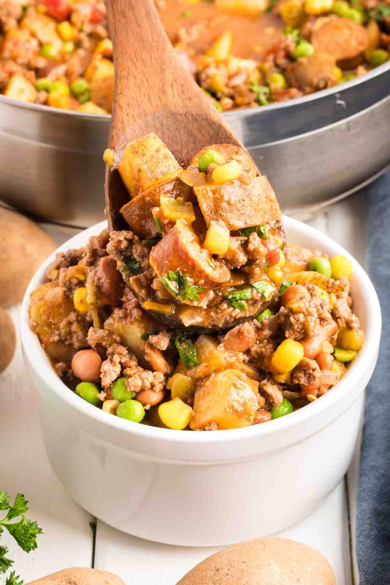 Hearty cowboy stew in a white bowl filled with ground beef, sausage, potatoes, corn, peas, and beans in a tomato-based broth, garnished with fresh parsley. Hostess At Heart