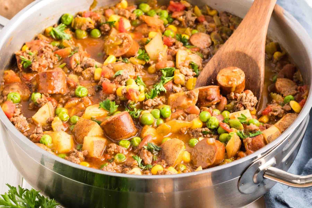 Closeup of a skillet filled with a Cowboy Stew made of ground beef, sausage, potatoes, corn, peas, and beans in a tomato-based broth,