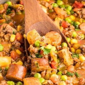 A wooden spoon lifting a serving of Cowboy Stew made of ground beef, sausage, potatoes, corn, peas, and beans in a tomato-based broth, garnished with fresh parsley. Hostess At Heart