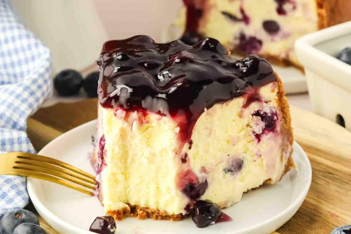 Close-up of blueberry cheesecake slice on white plate with blueberry sauce dripping down and fork a fork that's removed the first bite - Hostess At Heart