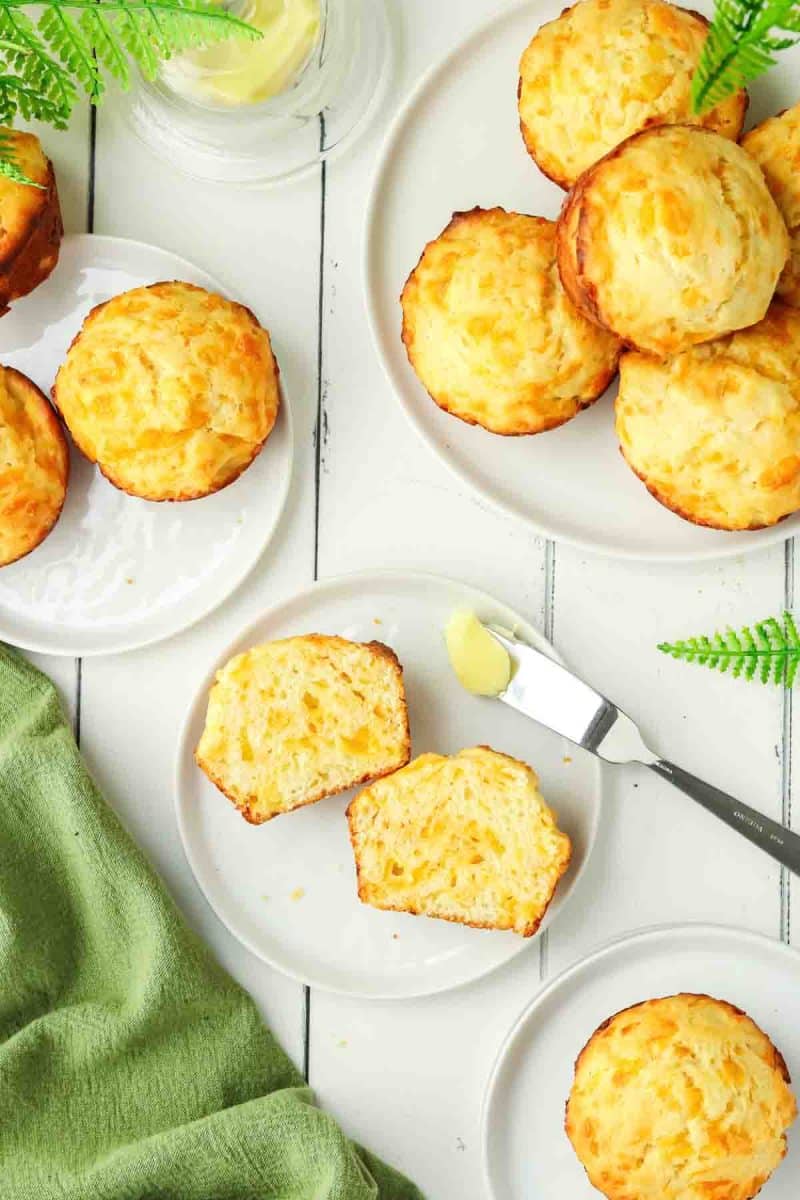 Top down view of plates filled with baked cheddar cheese muffins. The middle muffin is split open showing melted cheese inside - Hostess At Heart