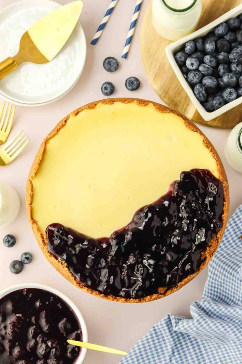 Blueberry cheesecake with glossy homemade blueberry sauce being spooned over half the top - Hostess At Heart