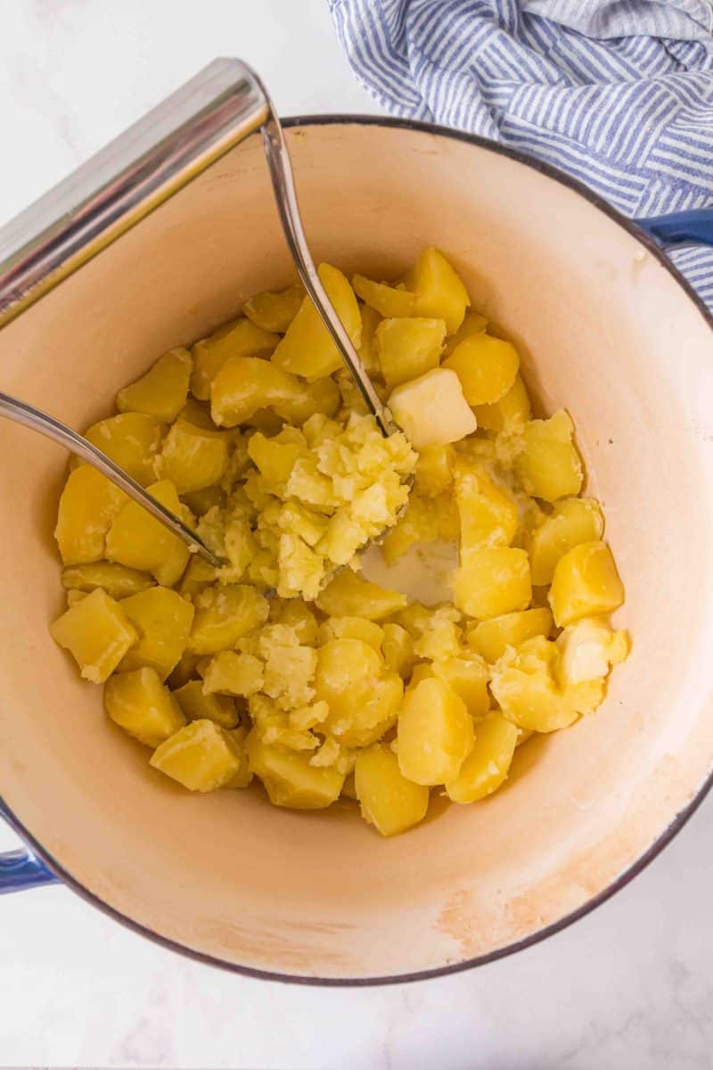 Mashing hot Yukon Gold potatoes with a hand masher in a pot - Hostess At Heart