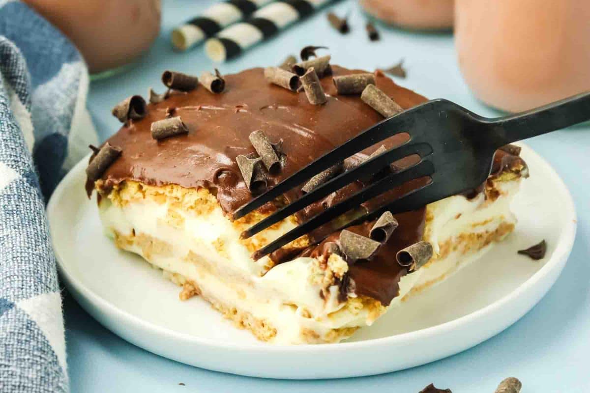 Slice of chocolate eclair cake on white plate showing layers of graham crackers, vanilla pudding, and chocolate frosting with a fork slicing through the piece - Hostess At Heart