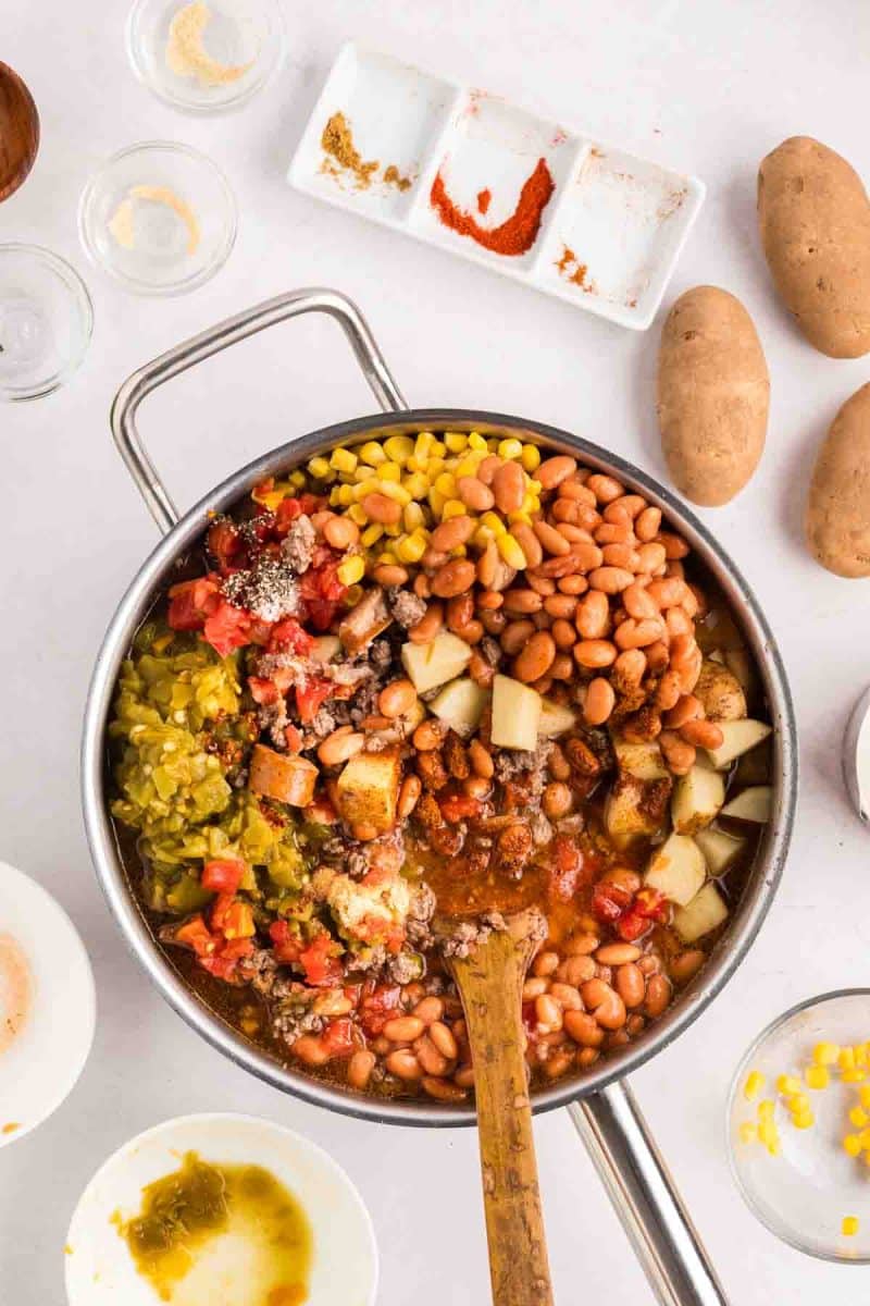 Ground beef, sausage, potatoes, corn, peas, and beans in a tomato-based broth being combined in a large skillet to make cowboy stew. Hostess At Heart