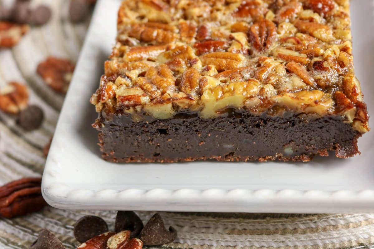 Sideview of a pecan pie brownie square on white plate showing caramelized pecan topping over a chocolate fudgy base. Hostess At Heart