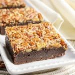 A sideview of a row of pecan pie brownie squares on white plate showing caramelized pecan topping - Hostess At Heart