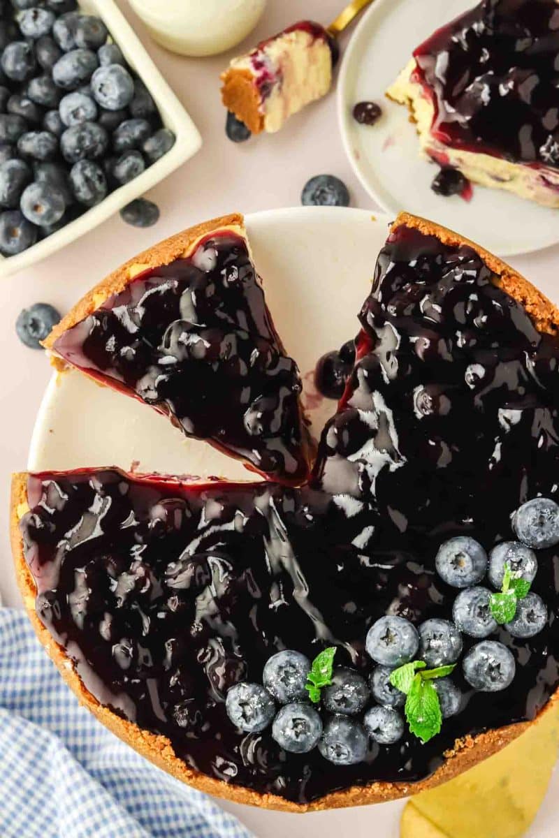 Top down view of a sliced blueberry cheesecake with glossy blueberry topping garnished with fresh blueberries and mint leaves - Hostess At Heart