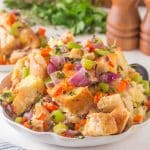 Angled view of baked sourdough stuffing in a white serving dish with golden brown bread, veggies and fresh herbs. Hostess At Heart