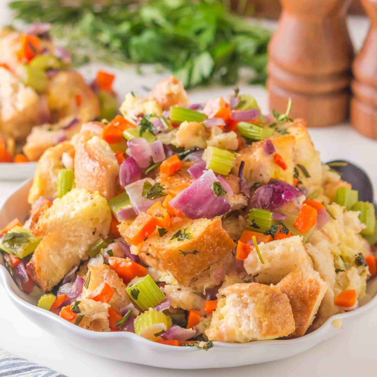Angled view of baked sourdough stuffing in a white serving dish with golden brown bread, veggies and fresh herbs. Hostess At Heart