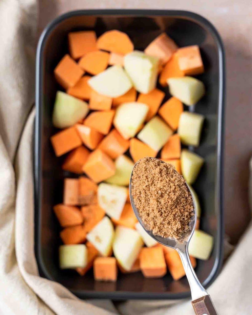 A casserole dish filled with chunks of sweet potatoes and apple. A spoon of a brown sugar mixture hovers over the pan. Hostess At Heart