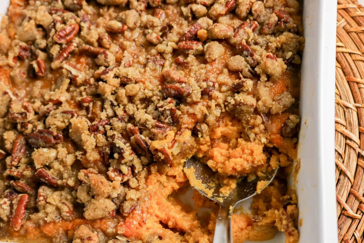 Top down view of a serving spoon scooping a portion of sweet potato casserole with pecan topping from a white baking dish - Hostess At Heart
