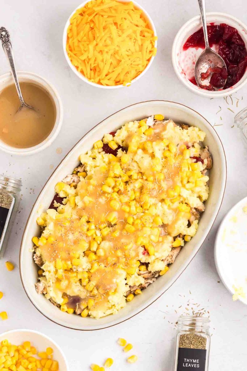 Gravy poured over a layer of corn, creamy mashed potatoes, cranberry sauce, and shredded turkey in baking dish - Hostess At Heart