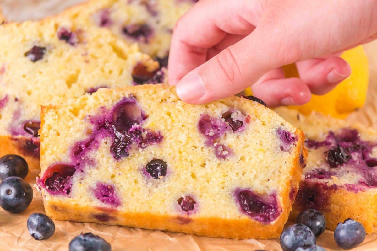 A hand reaching for a slice of lemon blueberry sourdough bread showing tender crumb and blueberries. Hostess at Heart